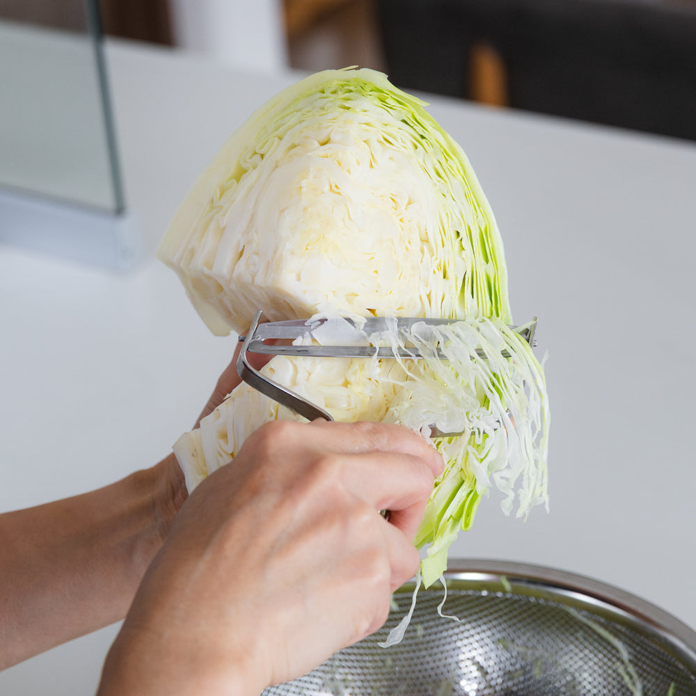 Person using a vegetable peeler to shred green cabbage into a metal bowl.