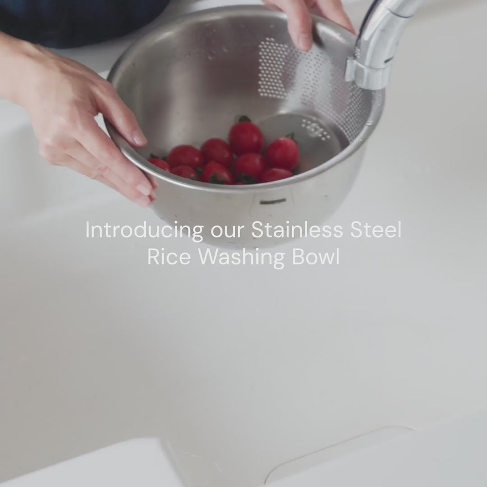 Hands rinse tomatos in a stainless steel rice washing bowl with built-in strainer under a kitchen faucet.