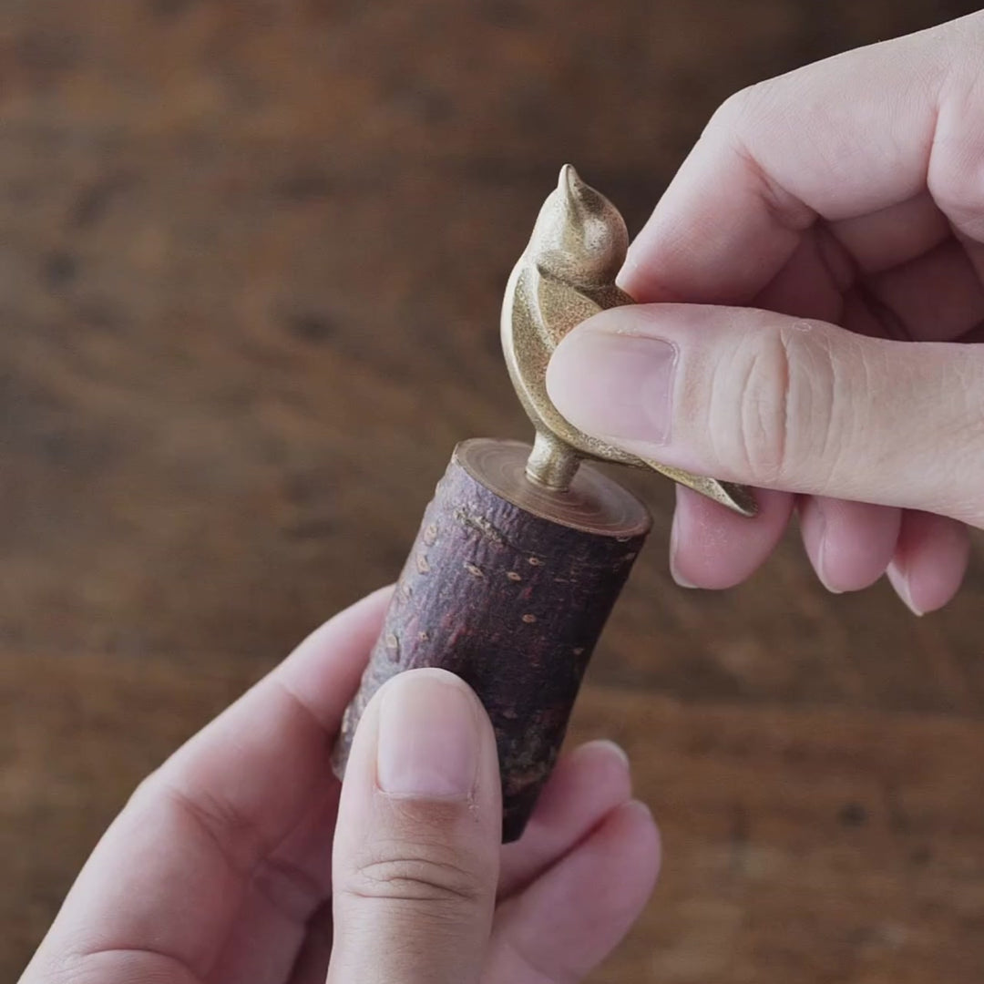A wooden base with a brass bird motif attached by a screw, producing birdsong when the screw is turned.