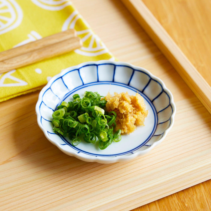White scalloped sauce dish with navy blue lines, holding chopped green onions and grated ginger, placed on a light wooden tray.