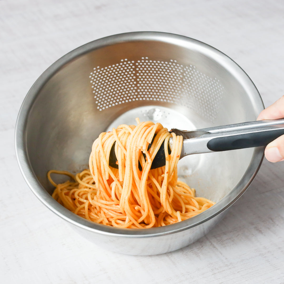 Tossing spaghetti in a stainless steel colander-style bowl, combining sauce and pasta for quick and even flavor coating.