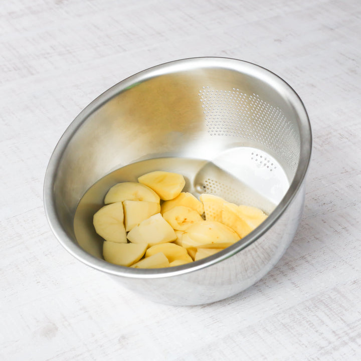 Chopped potatoes soaking in water inside a stainless steel bowl with built-in strainer holes, ready for boiling or cooking prep.
