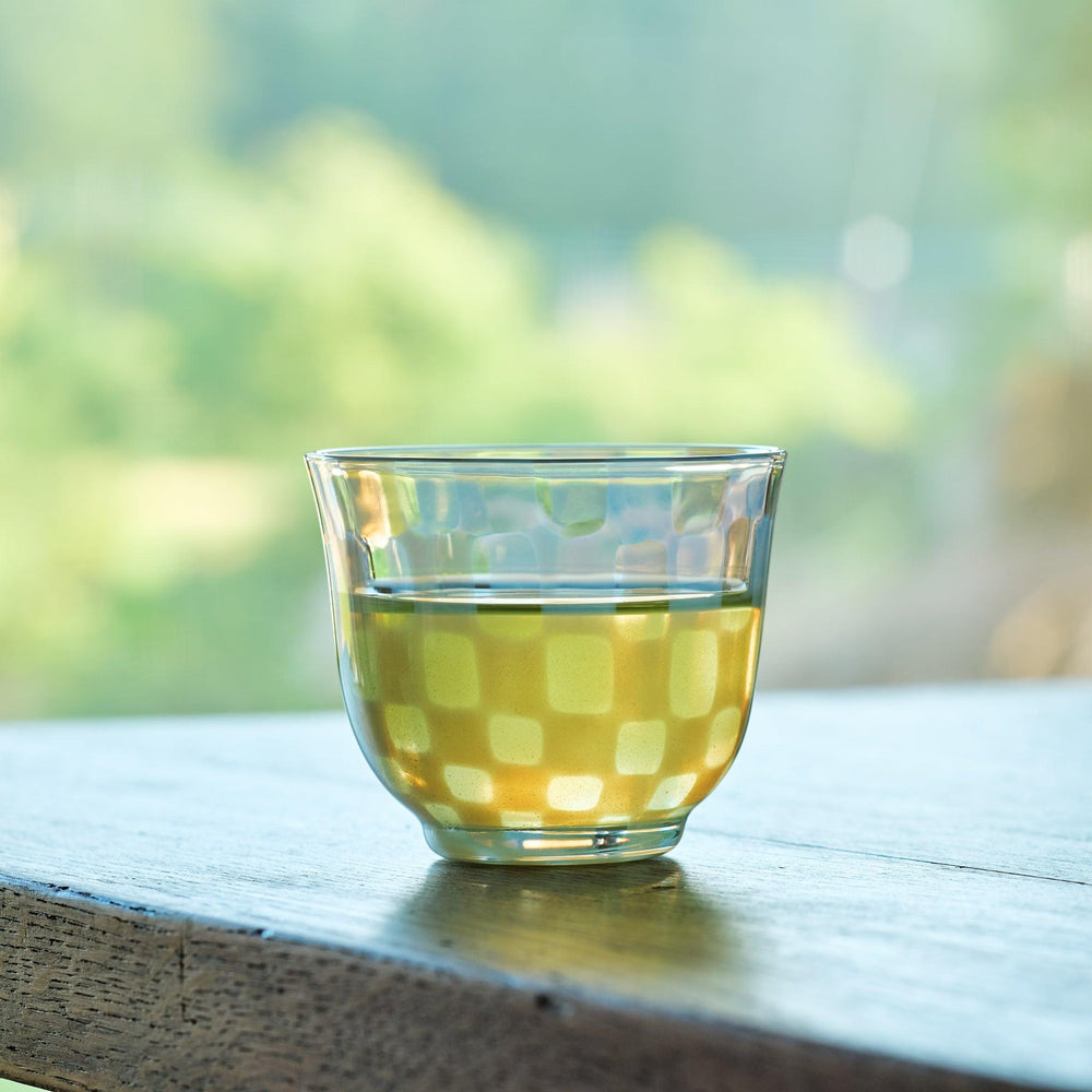 Vintage-style checkered glass tea cup filled with green tea, placed on a wooden table by a sunlit window.