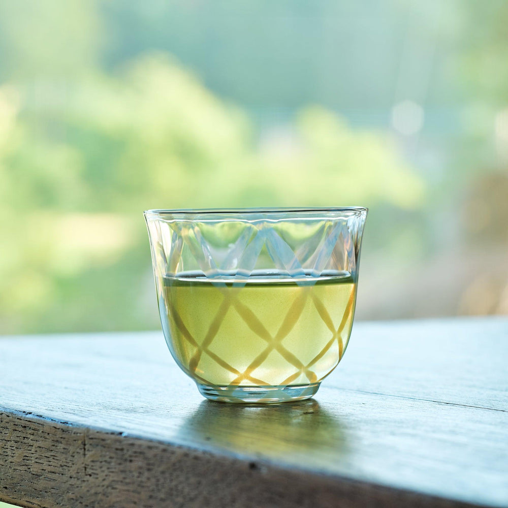 Vintage-style glass cup with a crisscross pattern and green tea, set on a wooden table by a sunlit window.