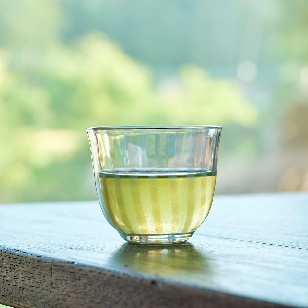 Japanese glass tea cup with translucent vertical stripes, filled with green tea and set by a sunlit window.