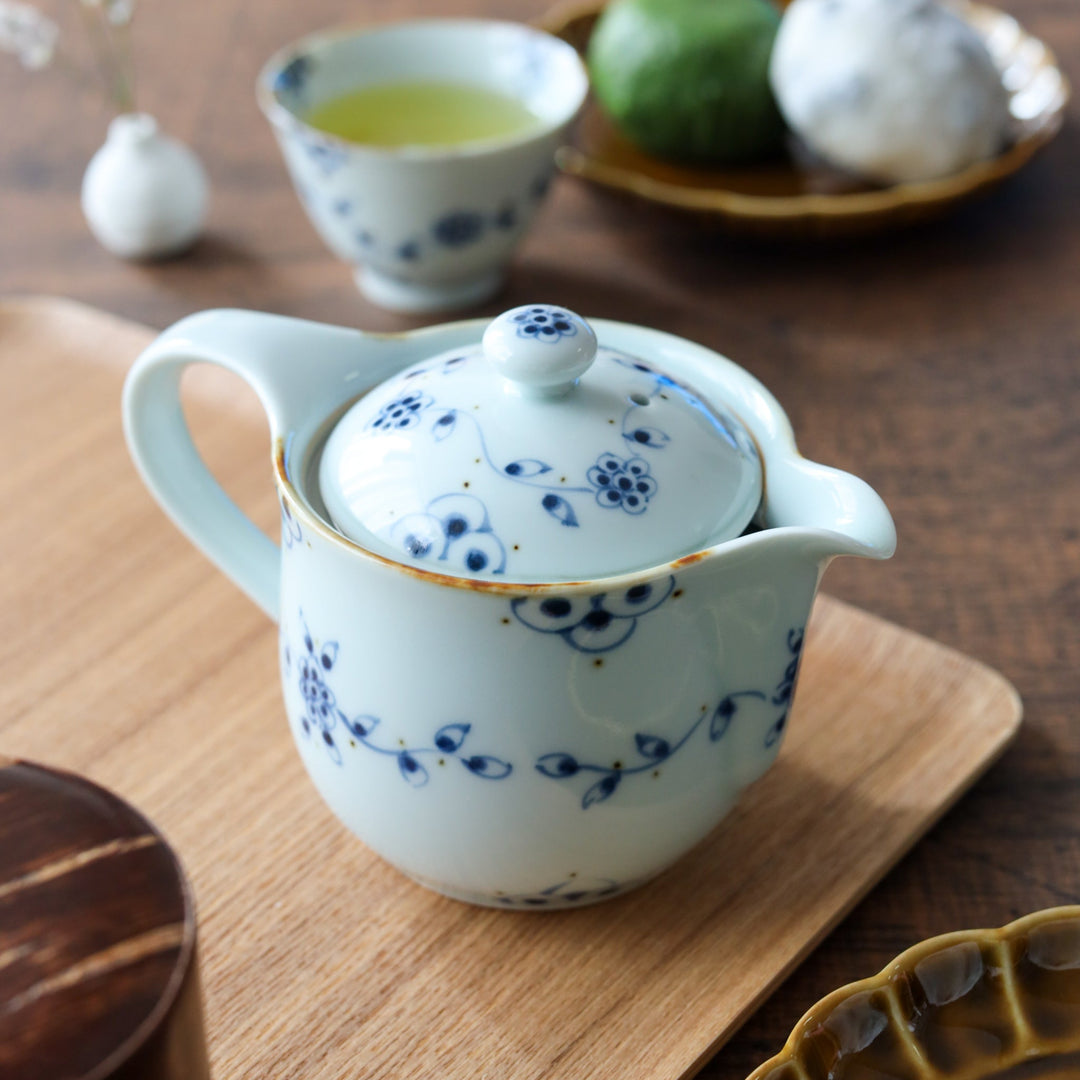 A hand-painted white porcelain teapot and two matching cups decorated with vintage blue vine motifs, displayed on an ash wood tray with Japanese sweets.