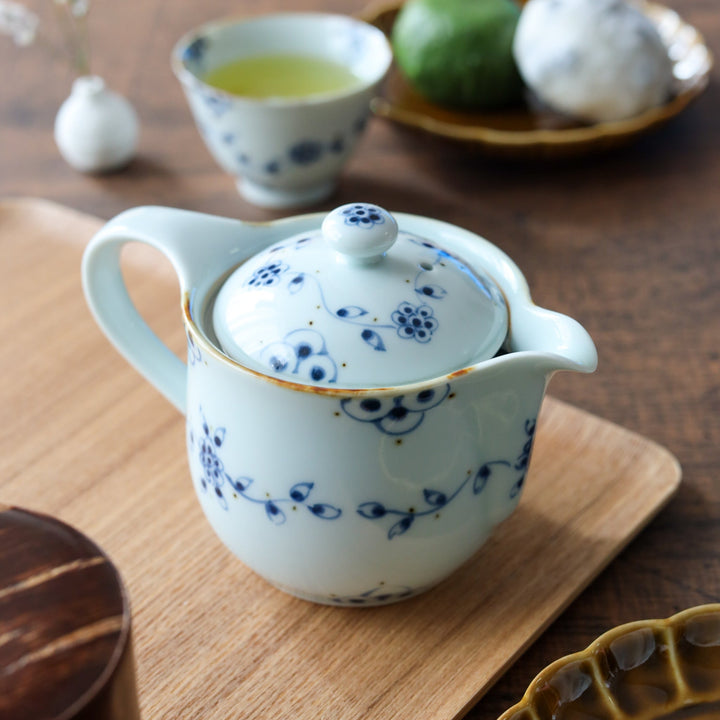 A hand-painted white porcelain teapot and two matching cups decorated with vintage blue vine motifs, displayed on an ash wood tray with Japanese sweets.
