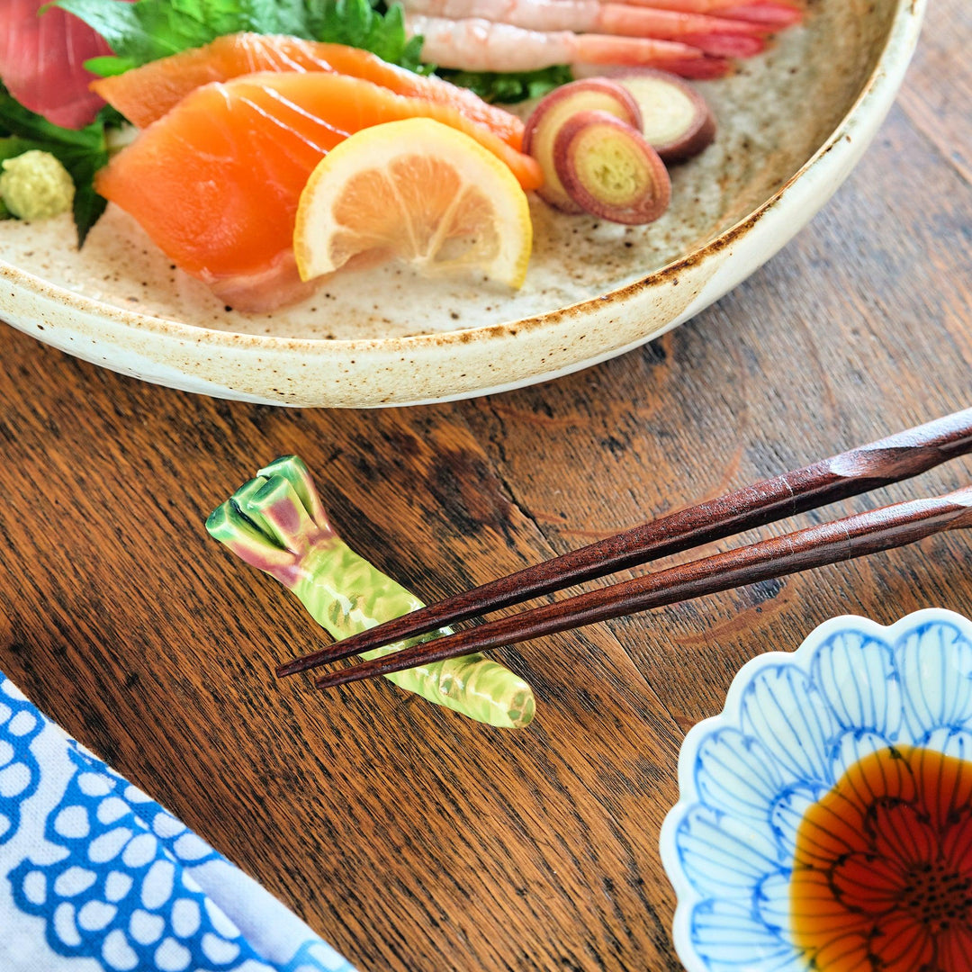 Wasabi-shaped ceramic chopstick rest in green and pink tones, holding wooden chopsticks on a rustic wooden table next to sashimi and a floral sauce dish.