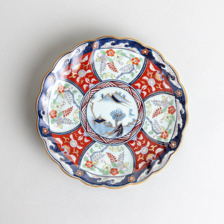 A scalloped porcelain bowl featuring an intricate Imari-style design with alternating red and blue patterns and a central landscape scene.