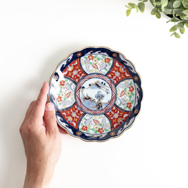 A scalloped porcelain bowl featuring an intricate Imari-style design with alternating red and blue patterns and a central landscape scene.