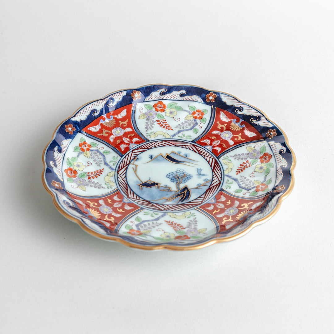 A scalloped porcelain bowl featuring an intricate Imari-style design with alternating red and blue patterns and a central landscape scene.